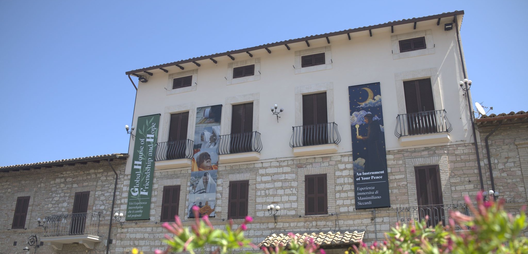 The facade of the Global House of Friendship and Hope in Santa Maria degli Angeli, Assisi, with banners and flowering plants in front