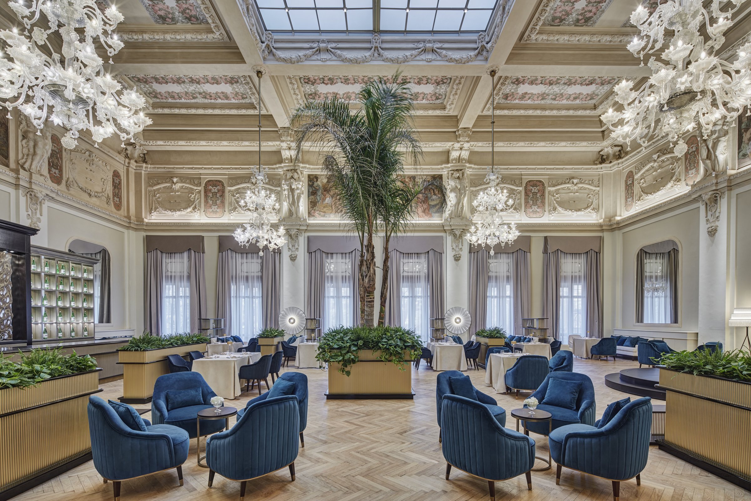 A frescoed hall with glass chandeliers, palms, and blue armchairs at Palazzo Fiuggi